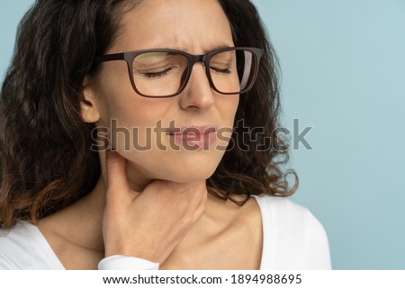 Woman having sore throat, tonsillitis, feeling sick, suffering from painful swallowing, angina, strong pain in throat, loss of voice, holding hand on her neck, isolated on studio blue background. Royalty-Free Stock Photo #1894988695