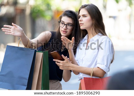 A woman carrying a shopping bag telling Give directions in the city for a woman hold a mobile phone. Young beautiful woman walking after successful shopping. Royalty-Free Stock Photo #1895006290
