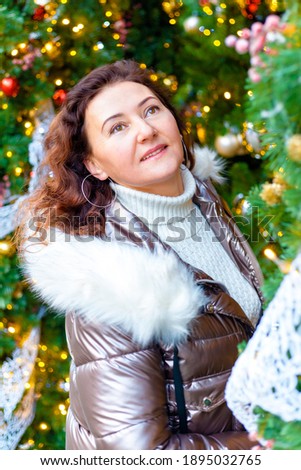 Cheerful European woman with long hair on a winter day against the background of bright Christmas lights and decorated Christmas tree.