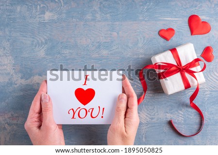Happy Valentine's Day greetings. Female hands hold a love message, valentine card with the inscription I LOVE YOU next to a gift with red hearts on a wooden grunge blue background