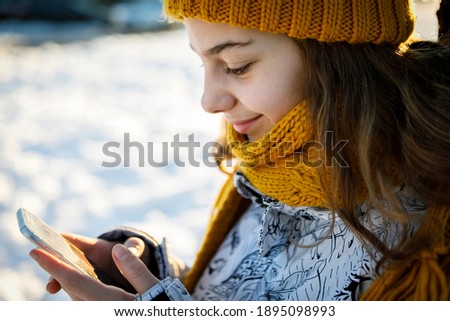 Closeup smiling teen girl in winter knit hat and warm scarf looking in smart phone over snowy background