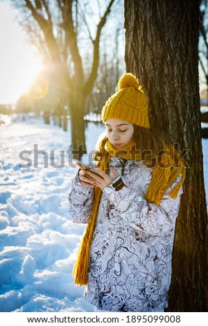 Closeup smiling teen girl in winter knit hat and warm scarf looking in smart phone over snowy background