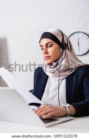 arabian translator in headset working with documents and typing on laptop on blurred foreground