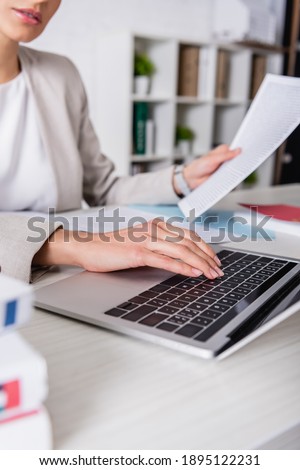 partial view of translator holding documents while typing on laptop, blurred foreground