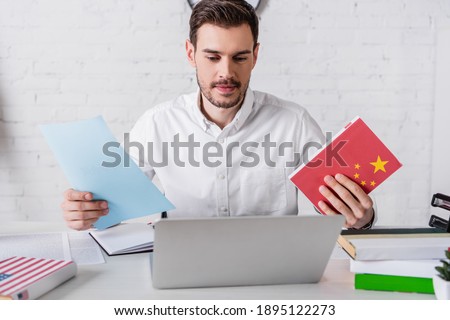 smiling interpreter holding chinese dictionary and document near laptop in translation agency