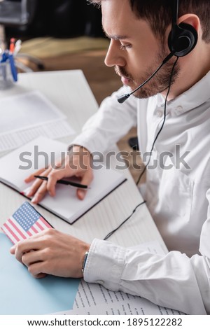 young interpreter in headset holding pen and digital translator with usa flag emblem, blurred background