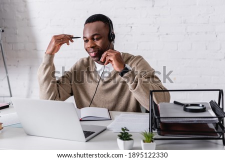 smiling african american interpreter in headset holding pen while working at laptop in translation agency