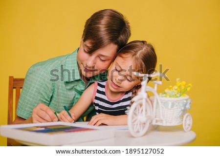 Children paint a picture while sitting at a table on a yellow background. Brother teaches little sister to draw with crayons. Studio photo