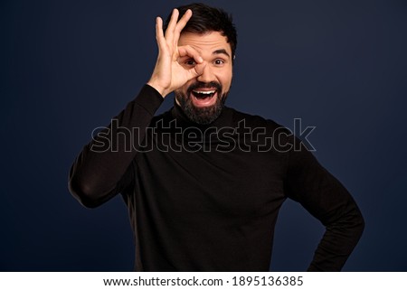 Cheeful, guy with beard wearingn black high neck sweater, showing okay, excellent, good gesture, make circle over eye and look through, with happy smile, agree, give recommendation, approve choice.