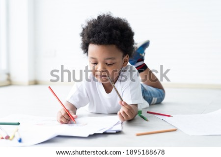 African American little boy lying and drawing on pages with colour pencils on floor at classroom. Kid learning by drawing. Education concept