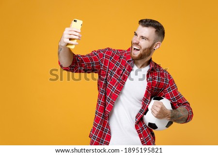 Joyful young man football fan in shirt cheer up support favorite team with soccer ball doing selfie shot on mobile phone clenching fists isolated on yellow background. People sport leisure concept