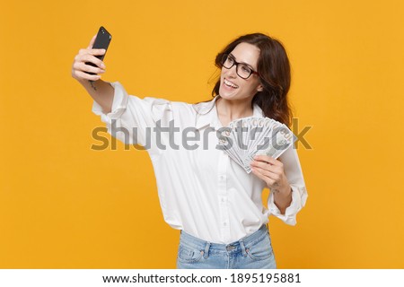Smiling business woman in white shirt glasses isolated on yellow background. Achievement career wealth business concept. Hold fan of cash money in dollar banknotes, doing selfie shot on mobile phone