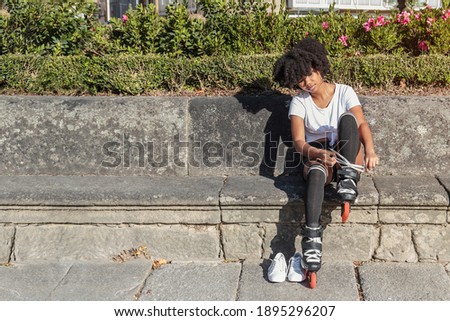 Young African American woman with afro hair is sitting putting on inline roller skates for outdoor skating training in park and playing sports on hot sunny day. Horizontal photography with copy space