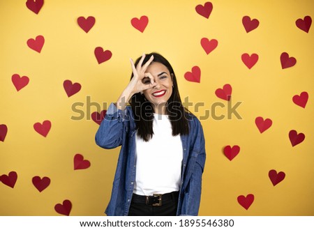Young caucasian woman over yellow background with red hearts doing ok gesture shocked with smiling face, eye looking through fingers