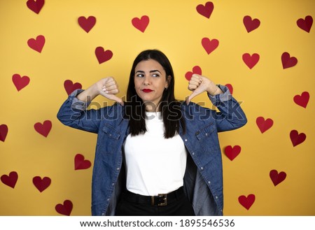 Young caucasian woman over yellow background with red hearts with angry face, negative sign showing dislike with thumb down