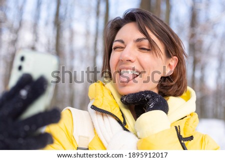 Young beautiful happy cheerful woman in the winter forest video blog, makes a selfie photo against the background of beautiful snow, smiles, dressed in warm clothes, yellow bright jacket