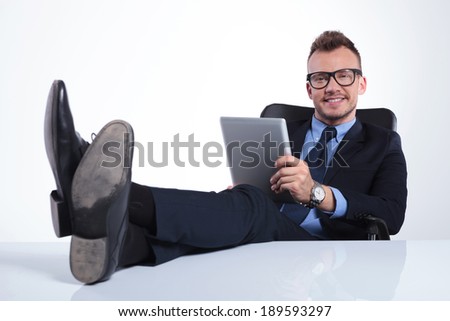 portrait of a young business man relaxing with his feet on the desk while holding his tablet and smiling for the camera. on gray background