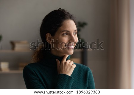 Smiling millennial Caucasian woman look in distance window dreaming or planning success. Happy young female thinking or pondering, visualize or imagine opportunity at home. Visualization concept.