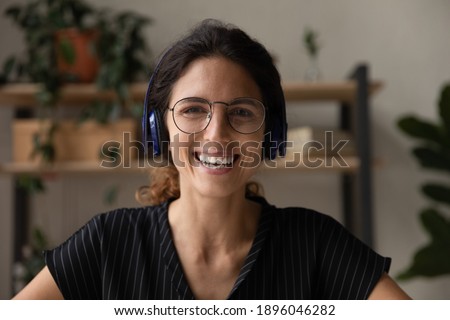 Close up headshot portrait of happy young Caucasian woman in glasses and headphones have webcam conference online. Profile picture of smiling female in earphones talk on video call or meeting.