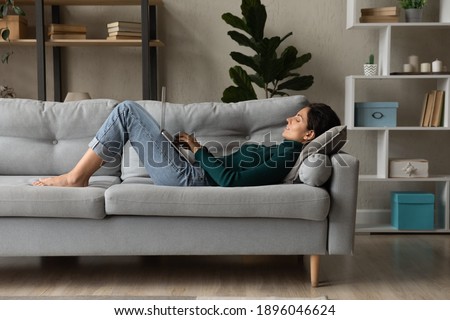 Young Caucasian woman lying on couch in living room work online on laptop gadget. Millennial female relax rest on sofa at home look at computer screen text message on device. Communication concept. Royalty-Free Stock Photo #1896046624