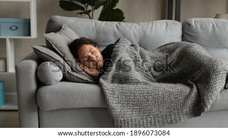 Happy millennial Caucasian woman lying under blanket on cozy couch at home sleeping or dreaming. Smiling calm young female relax rest on sofa in living room, relieve negative emotions daydreaming. Royalty-Free Stock Photo #1896073084