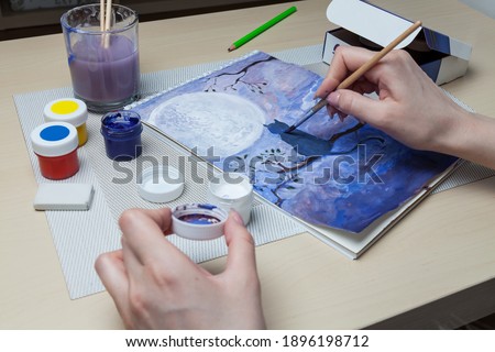 A close-up of female hands over a painted picture holding a brush and a cover for watercolors on which the colors are mixed next to the eraser and a simple pencil. A hobby of art.