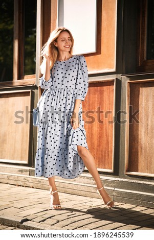 Beautiful young woman in stylish light blue polka dot dress with handbag on city street Royalty-Free Stock Photo #1896245539