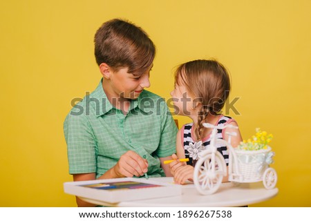 Children paint a picture while sitting at a table on a yellow background. Brother teaches little sister to draw with crayons. Studio photo