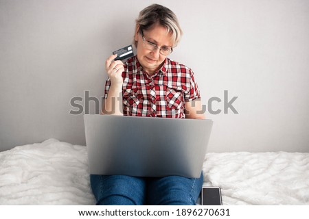 Attractive middle-aged woman with glasses sitting with laptop and credit card in light room, shopping online. Cozy way of shopping
