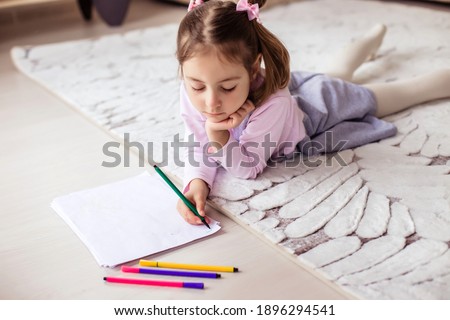A little girl lies on a light carpet on the floor and draws with markers. Classes with children at home