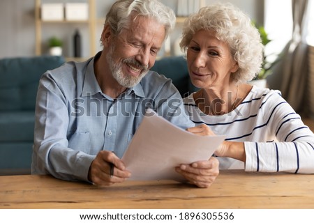 Happy senior family couple looking at paper sheet, reading letter with pleasant news. Smiling middle aged husband and wife holding correspondence mail or medical insurance contract together at home. Royalty-Free Stock Photo #1896305536