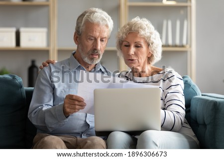 Happy elderly senior family couple reading paper correspondence or mails, sitting on sofa with computer on laps. Smiling mature spouses managing financial affairs indoors using laptop in living room. Royalty-Free Stock Photo #1896306673