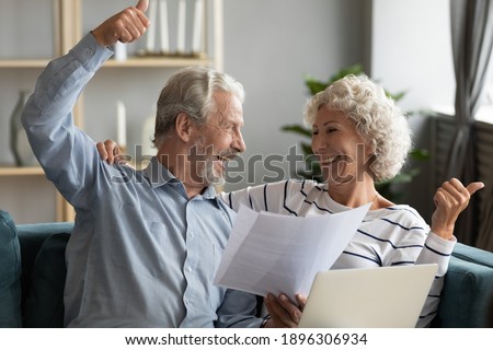 Overjoyed emotional middle aged mature family couple holding paper financial documents in hands, celebrating getting bank loan approval notification, tax refunds or reading good news together at home. Royalty-Free Stock Photo #1896306934