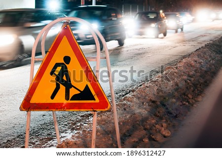 Triangular warning sign road works stands on the snowdrift roadside in the background cars pass with their headlights on at night after snowfall