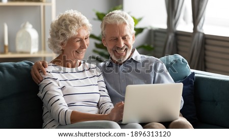 Happy middle aged older retired family couple using computer applications, having fun web surfing internet, shopping online or communicating distantly relaxing together on cozy sofa in living room.