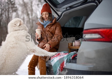 Young woman with her white dog drink a coffee in car trunk near a snowy forest, traveling by car during winter holidays. High quality photo