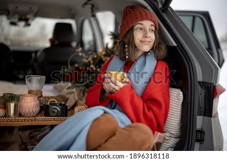 Woman with hot tea sitting in car trunk and look at the forest, traveling by car during winter holidays. High quality photo
