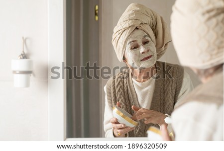 Elderly woman with towel on her head smiles and applies cleansing mask on her face. Happy pensioner looks after her appearance, puts on a white natural mask with collagen, tighten and rejuvenate face.