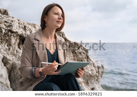 Attractive concentrated short haired woman with sketchbook and pencil sitting on old rock near tranquil sea on nasty day