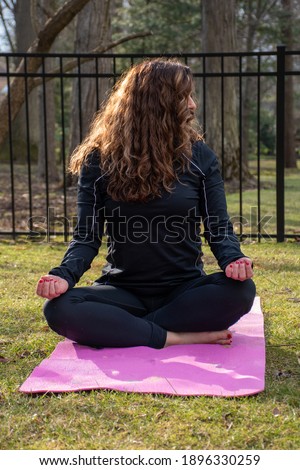 woman with curly hair over her face nose peeking out unrecognizable looking to the side in yoga crossed legs wearing black on pink mat in grass outside