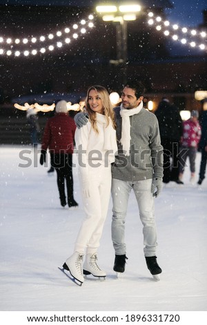 Happy stylish couple of lovers having fun, looking away, standing at ice skating rink outdoor. St. Valentines Day, Winter leisure, New Year holidays concept.