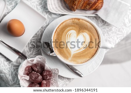 still life flat lay of an Italian breakfast: hard-boiled egg, cappuccino with heart foam, marron glaces, croissants. breakfast presented on a gray table runner and matching napkins Royalty-Free Stock Photo #1896344191