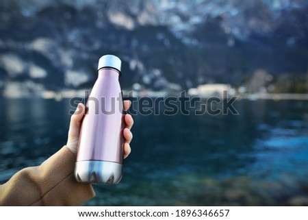 Close-up of female hand holding reusable, steel thermo shiny bottle for water, on the background of clear water of a lake with a turquoise hue. Copy space concept.  Royalty-Free Stock Photo #1896346657