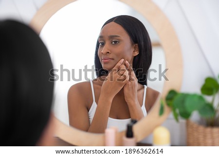 Acne treatment, squeezing, spot, skin care, pressing on skin problem face. Sad young african american woman looking in mirror and removing pimple from face at home in interior bedroom in morning