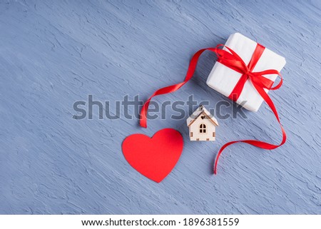 A symbol of love, family, relationships. Gift in white paper with a red ribbon, red heart shape and copy of the house on a stylish gray background, copy space. Valentine's Day, Family Day