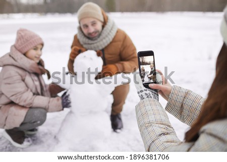 Portrait of young woman taking photo of father and daughter building snowman together, focus on smartphone screen, copy space