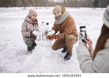 Portrait of young woman taking photo of father and daughter building snowman together while enjoying winter outdoors, copy space