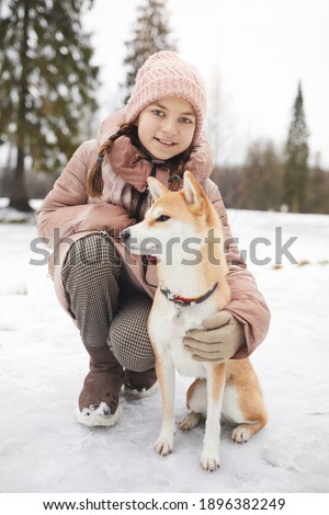 Vertical full length portrait of smiling girl posing with pet dog and looking at camera while enjoying walk outdoors in winter forest