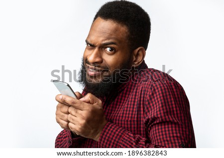 Phone Fraud. Suspicious Black Man Using Smartphone Looking At Camera Posing Over White Studio Background. Telephone Phreaking And Cellphone Scam Concept Royalty-Free Stock Photo #1896382843