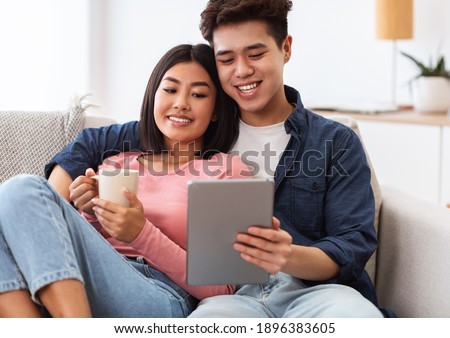 Asian Boyfriend And Girlfriend Using Tablet Computer Watching Movie Online Embracing Sitting On Sofa At Home Relaxing On Weekend Morning. Modern Gadgets, Chinese Family Lifestyle